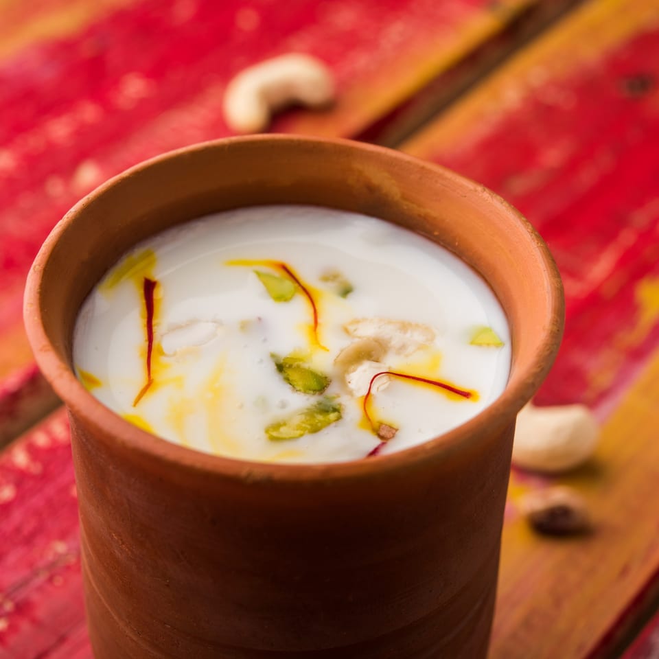 Indian lassi in a clay cup, garnished with saffron, pistachios, and cashews on a red wooden surface.